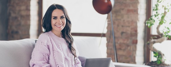 Junge Frau mit fliederfarbenen Pulli sitzt mit Laptop auf der Couch und lächelt