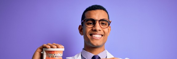 Zahnarzt zeigt auf ein Gebiss (auf lila Hintergrund)